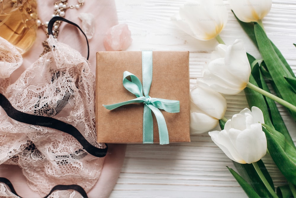 Boîte-cadeau élégante et bijoux de lingerie en dentelle et parfum sur tissu doux et tulipes sur fond rustique blanc. Essentiel pour femme laïque plate pour des vacances. Fête des mères ou anniversaire des femmes