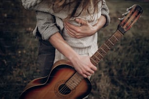 Pareja de músicos hipster abrazando en el campo, hombre guapo abrazando a la mujer gitana en vestido blanco, primer plano de la guitarra
