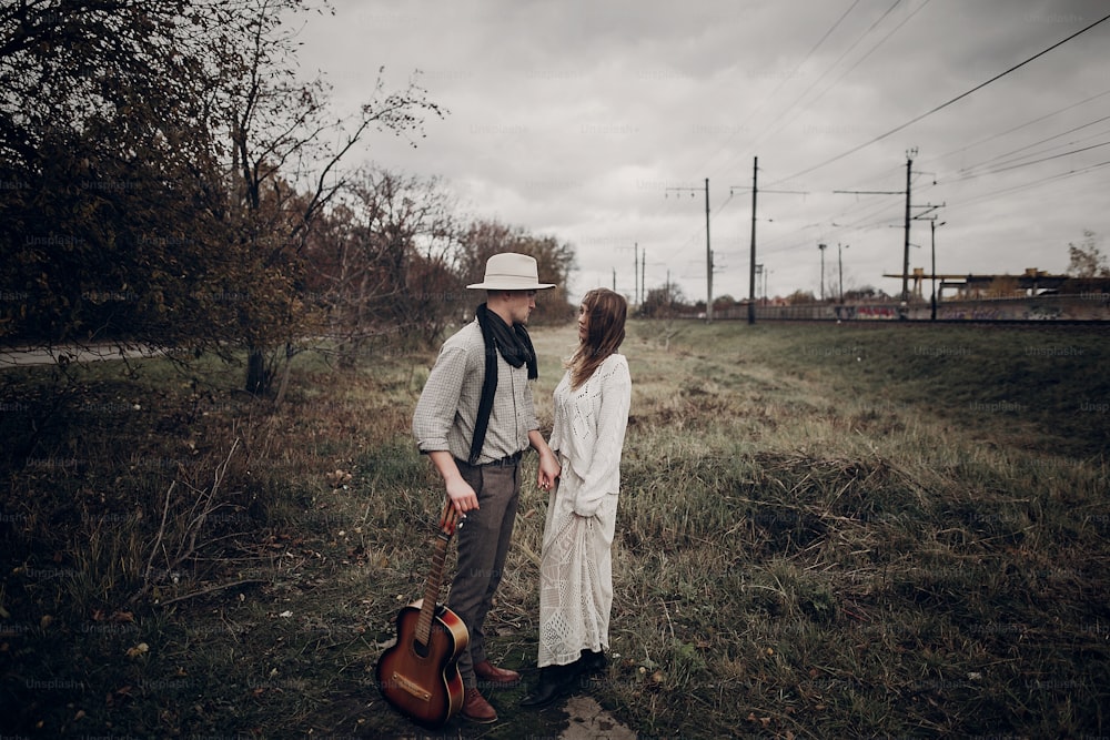 stylish hipster couple posing in windy field. boho gypsy woman and man with guitar in windy field. atmospheric motion moment. fashionable look. rustic wedding concept