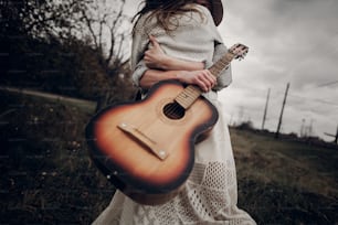 Hipster musician couple hugging in field, handsome man embracing gypsy woman in white dress, guitar closeup