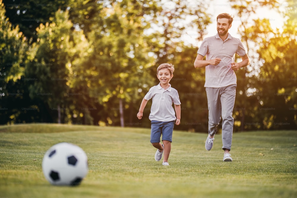 Handsome dad with his little cute sun are having fun and playing football on green grassy lawn