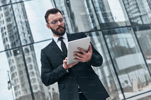 Good looking young man in full suit using digital tablet while standing outdoors