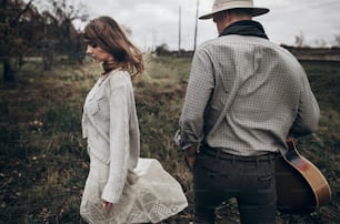 stylish hipster couple posing. man in hat with guitar and his boho woman in knitted sweater in windy field. atmospheric sensual moment. fashionable look. rustic wedding concept