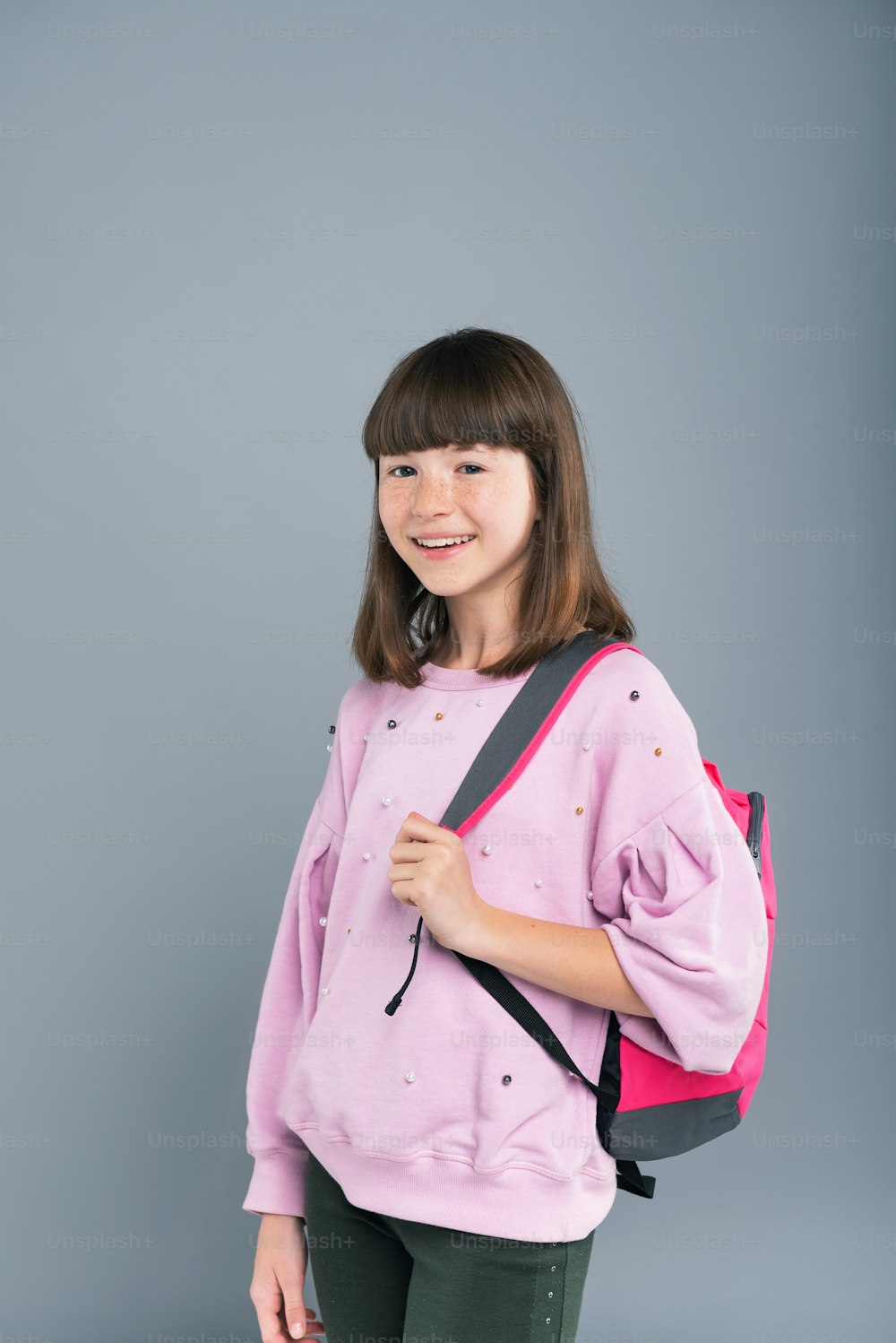 Bereit für die Schule. Fröhliches Teenager-Mädchen mit einem Rucksack auf der Schulter und lächelt in die Kamera, während es isoliert auf einem blaugrauen Hintergrund posiert
