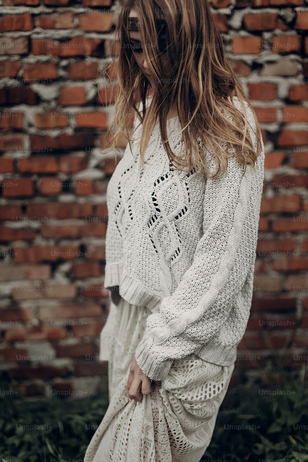 Beautiful brunette hipster woman in white boho sweater and dress posing near old wall