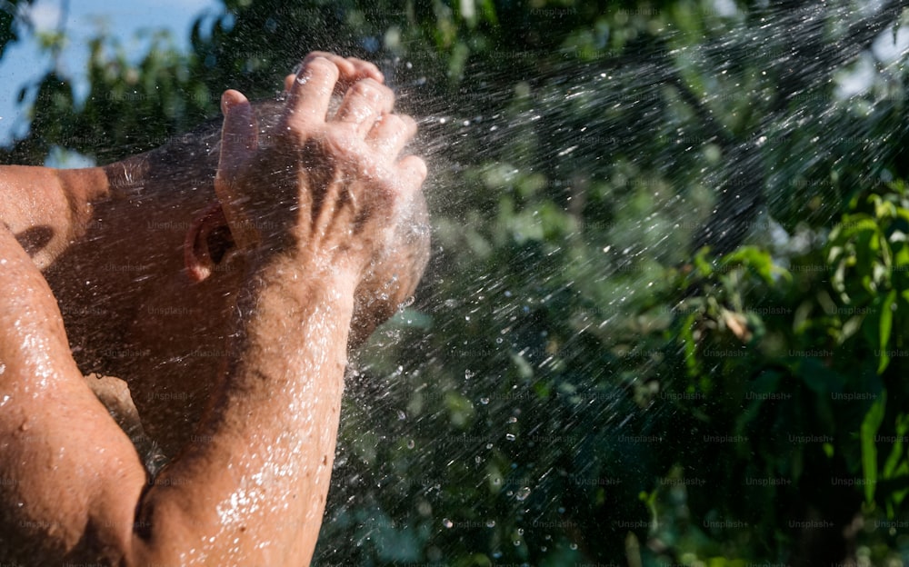L'uomo adulto fa la doccia sotto un tubo da giardino in un giardino verde estivo. Pittoresco cortile estivo