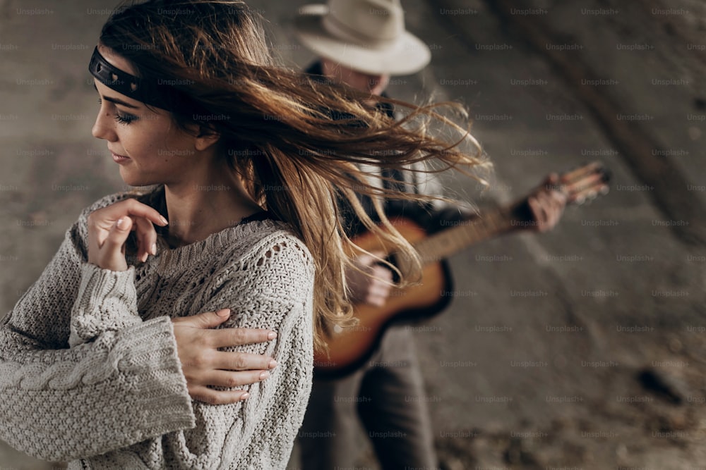세련된 힙스터 커플 포즈. 니트 스웨터를 입은 보헤미안 여자를 위해 기타를 연주하는 모자를 쓴 남자. 분위기 있는 관능적인 순간. 소박한 패셔너블한 룩.
