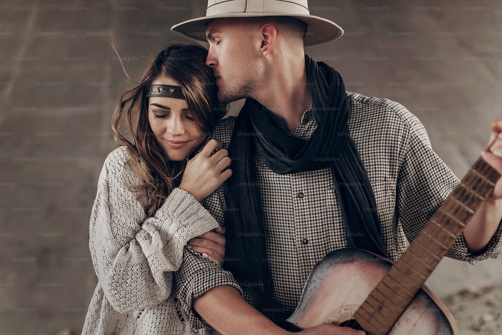 Stilvolles Hipster-Paar umarmt sich sanft. Mann mit Hut spielt Gitarre für seine Boho-Frau im Strickpullover. atmosphärischer sinnlicher Moment. rustikaler modischer Look.
