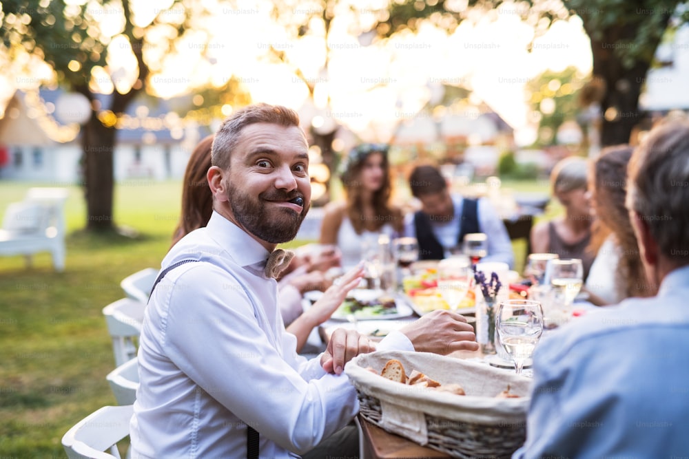 Guests sitting at the table and eating at the wedding reception outside in the backyard. A mature man making funny faces.