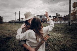 Sensual hipster couple, gypsy woman in boho clothes and musician with guitar wave at passing train