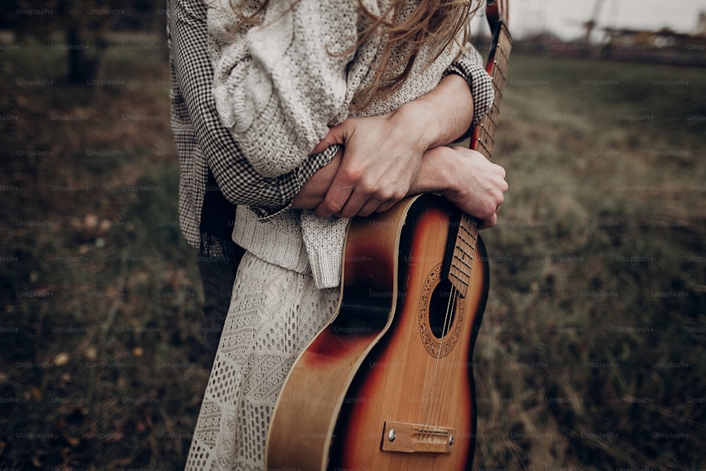 Pareja de músicos hipster abrazando en el campo, hombre guapo abrazando a la mujer gitana en vestido blanco, primer plano de la guitarra