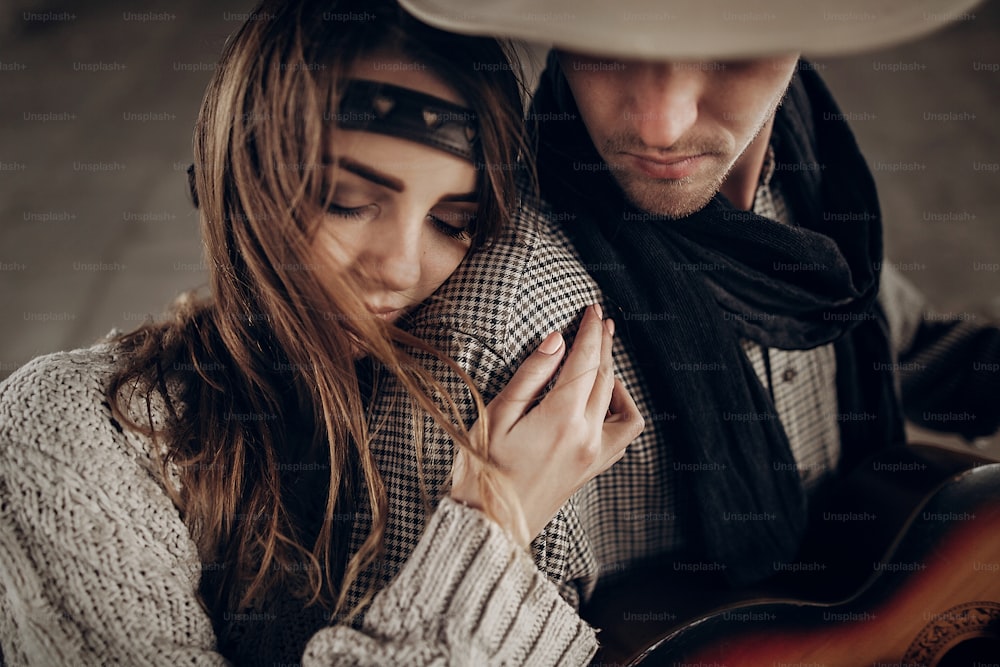 Romantic brunette hipster girl in boho clothes hugging handsome cowboy musician with guitar and a scarf closeup