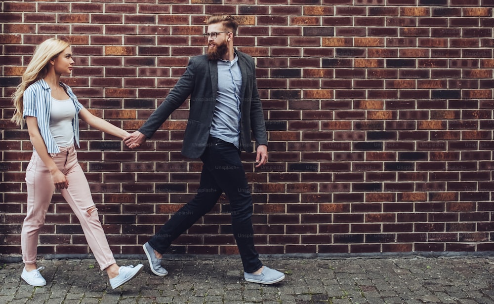 Stylish young couple is spending time together outdoors. Attractive woman and handsome bearded man are walking on brick background and holding hands.