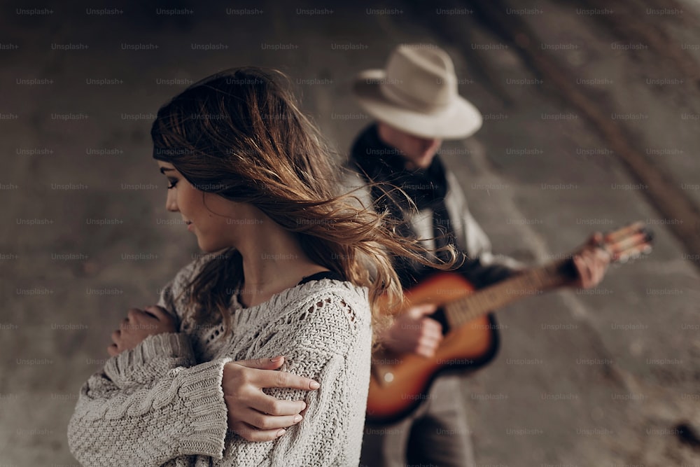 Beautiful sensual indie girl in hipster boho clothes posing in front of handsome cowboy guitar player outdoors