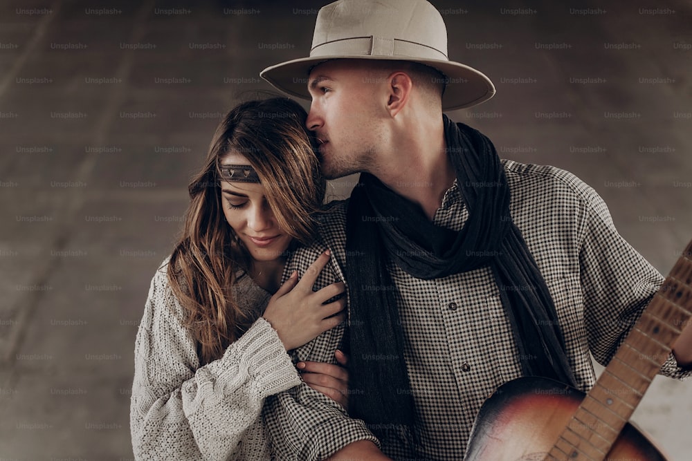 Hübscher Cowboy-Mann mit einer Gitarre, der eine schöne Indie-Frau küsst, während er im Freien posiert