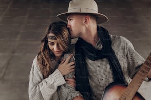 Hübscher Cowboy-Mann mit einer Gitarre, der eine schöne Indie-Frau küsst, während er im Freien posiert
