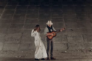 Stylish gypsy woman in boho clothes dancing near handsome man, cowboy musician with guitar