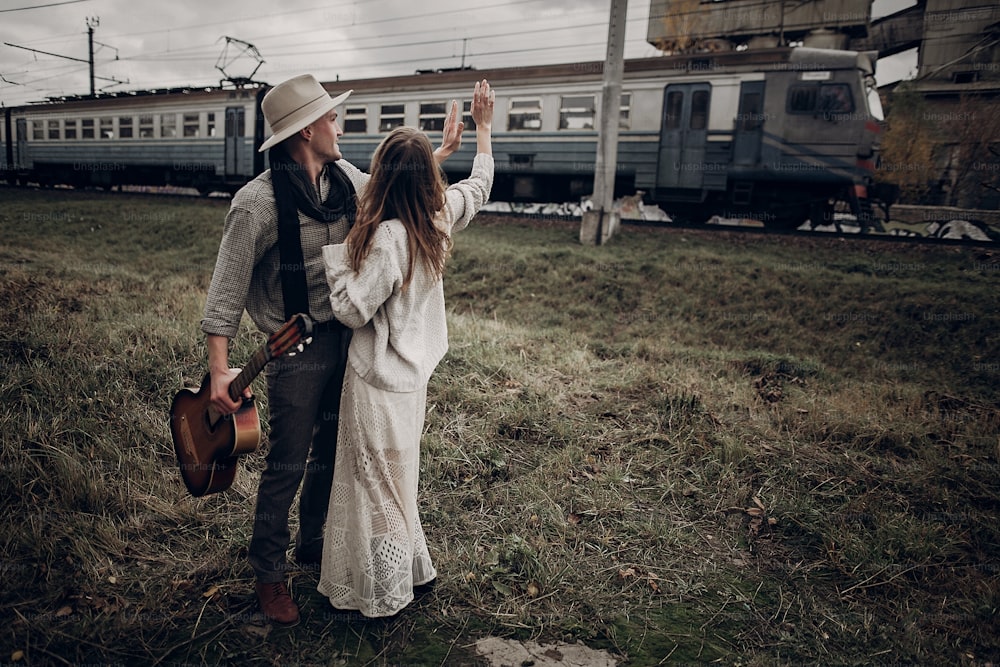 Sensual hipster couple, gypsy woman in boho clothes and musician with guitar wave at passing train