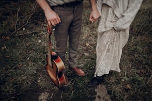 rustic wedding concept. boho gypsy woman and man with guitar posing in windy field. stylish hipster couple holding hands. atmospheric sensual moment. fashionable look.
