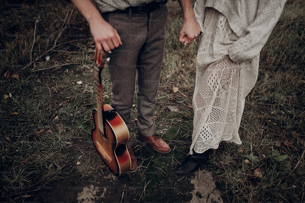 rustic wedding concept. boho gypsy woman and man with guitar posing in windy field. stylish hipster couple holding hands. atmospheric sensual moment. fashionable look.