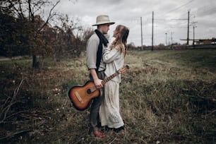 Rustikales Hochzeitskonzept. Boho Zigeunerin und Mann mit Gitarre posieren im windigen Feld. Stilvolles Hipster-Paar umarmt sich. atmosphärischer sinnlicher Moment. Modischer Look.