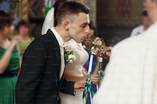 幸せな精神的なカップル、スタイリッシュな新郎、白いドレスを着た美しいブルネットの花嫁は、黄金の王冠の戴冠式中に結婚式の教会の式典でろうそくを保持しています