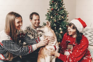 merry christmas and happy new year concept. stylish hipster family in festive sweaters playing and smiling with cute dog at christmas tree lights. happy holidays. atmospheric emotional moments