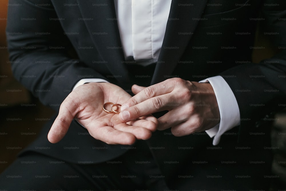 Wedding rings on palm hand. Groom in stylish suit holding golden wedding rings in hands, sitting in the room. marriage or divorce concept