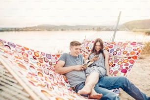 happy couple looking at phones and smiling, relaxing in hammock in sunset light on the beach. stylish hipsters having fun, and resting on summer vacation. comunication and social media concept