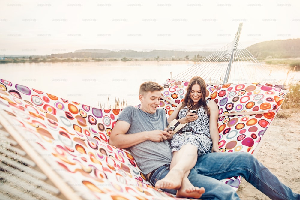 happy couple looking at phones and smiling, relaxing in hammock in sunset light on the beach. stylish hipsters having fun, and resting on summer vacation. comunication and social media concept