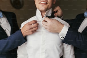 Groomsmen helping groom getting ready in the morning in the room. man hands putting on jacket and bow tie, standing at window light in hotel. preparation for wedding day