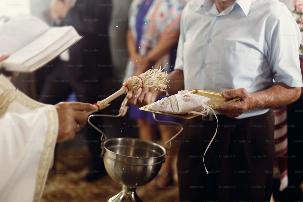 Prêtre chrétien orthodoxe arrosant des alliances en or sur un oreiller blanc avec de l’eau bénite, rituel religieux sacré à l’église lors de la cérémonie de mariage