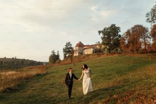 gorgeous bride and stylish groom walking holding hands in soft evening light at sunset near castle. sensual romantic moment of beautiful wedding couple outdoors