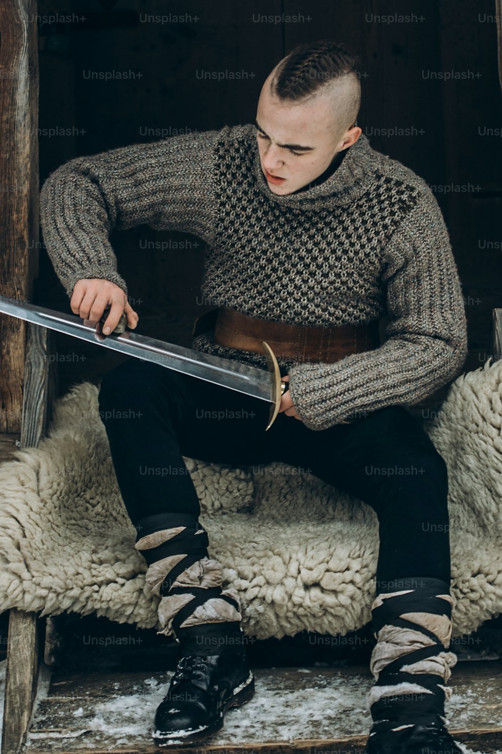 Portrait d’un guerrier viking fort aiguisant son épée, cosplay viking scandinave, guerrier médiéval avec une épée en acier assis sur une peau de loup dans les bois d’hiver, concept de préparation au combat