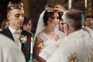Couple spirituel heureux, marié élégant et belle mariée brune en robe blanche tenant des bougies lors de la cérémonie de mariage à l’église lors du couronnement de la couronne d’or