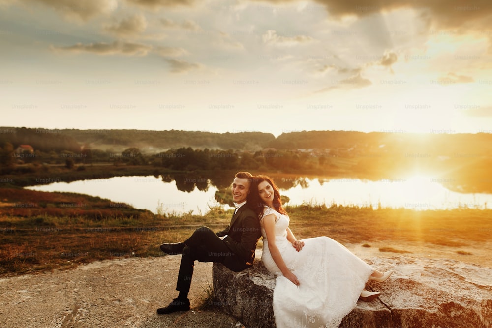 happy wedding couple sitting back to back and smiling in soft evening light at sunset. emotional moment of gorgeous bride and stylish groom in amazing sunset outdoors