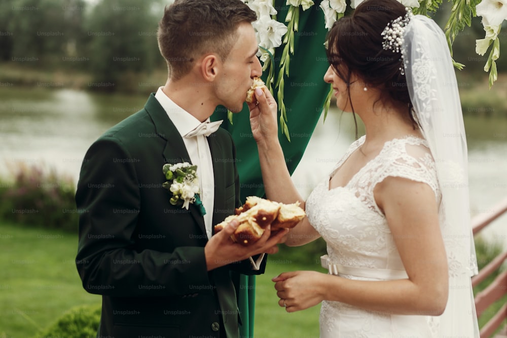 Heureux couple de jeunes mariés rompant le pain lors d’une cérémonie de mariage, beau marié et mariée souriante mangeant du pain près de l’allée du mariage pendant la tradition slave en plein air
