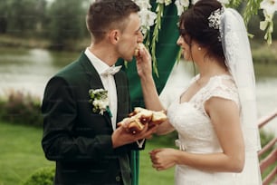 Happy newlywed couple breaking bread at wedding ceremony, handsome groom and smiling bride eating bread near wedding aisle during slavic tradition outdoors