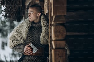 Ancient viking warrior with steel axe posing near wooden hut in scandinavian winter forest, thor viking cosplay, traditional norway costume, handsome man with mohawk hairstyle standing outdoors