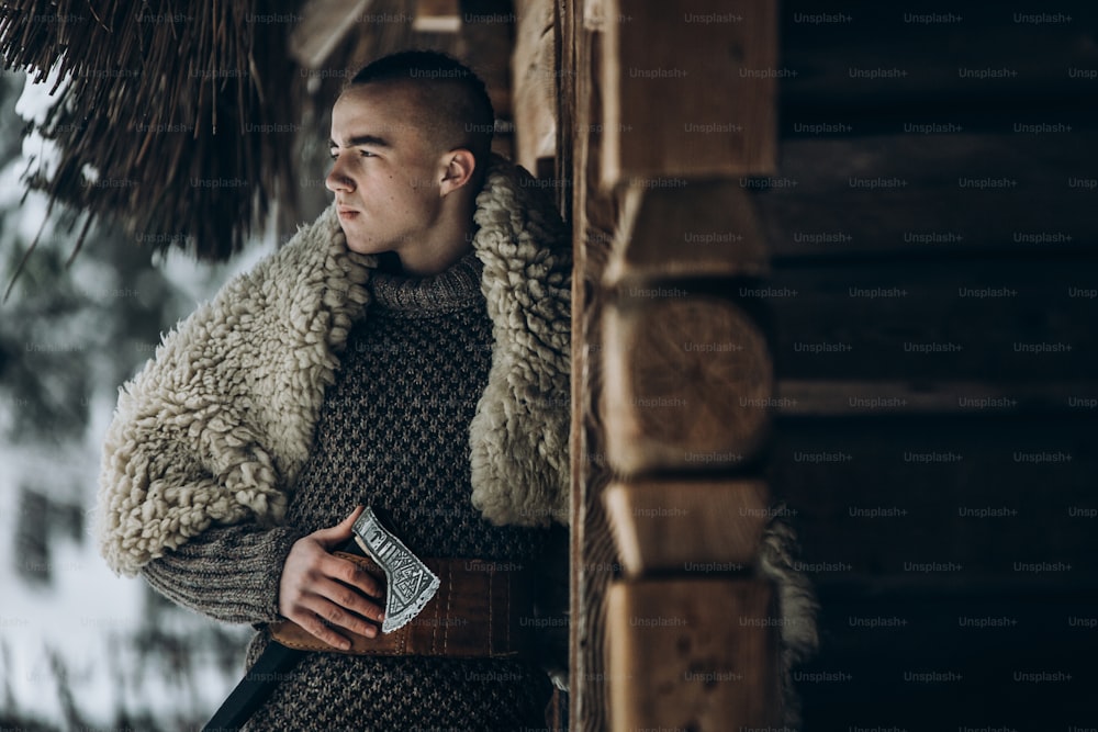 Antiguo guerrero vikingo con hacha de acero posando cerca de una cabaña de madera en el bosque invernal escandinavo, cosplay vikingo de Thor, traje tradicional de Noruega, hombre guapo con peinado mohawk de pie al aire libre