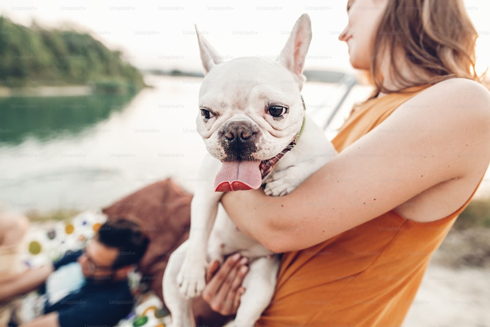 Mujer feliz sosteniendo bulldog lindo con la lengua fuera, marido en una hamaca en el fondo, chica hipster con su mascota
