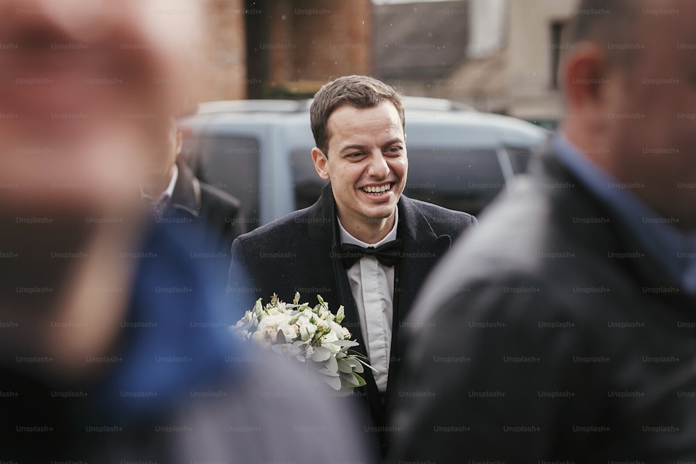 Stylish groom in suit  holding wedding bouquet and walking from car to bride among groomsmen and friends.