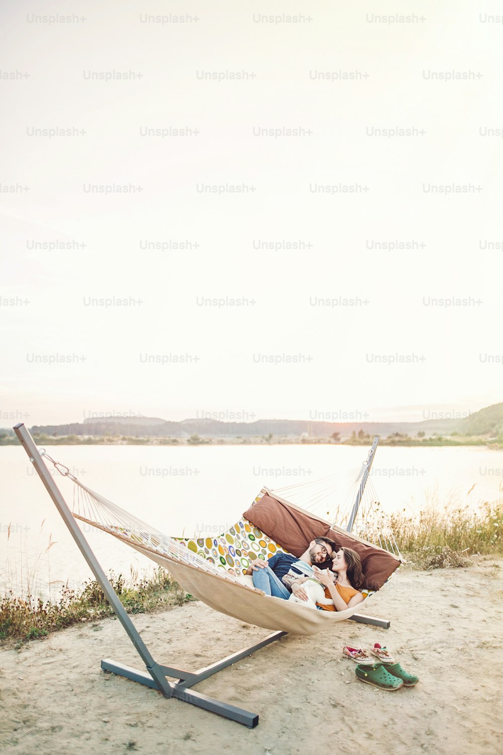 happy hipster couple with bulldog relaxing in hammock on the beach in sunset light, summer vacation. stylish family with dog cuddling and having fun, cute moments in summer evening