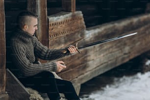 Valiente guerrero sosteniendo la espada cerca del edificio histórico del castillo de madera en los bosques de invierno en Escandinavia, vikingo guapo con mohawk posando con arma antes de la batalla, concepto de cosplay de fantasía