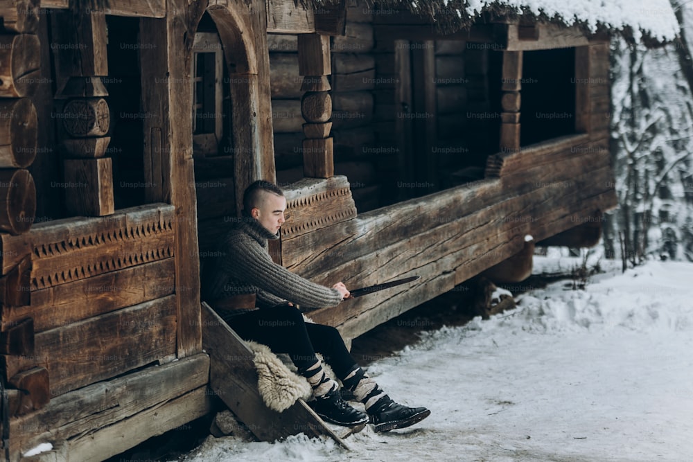Starker Wikingerkrieger, der sein Schwert schärft, während er in der Nähe einer alten Holzburg sitzt, skandinavischer Ritter mit Waffe im Wikingerkostüm, historisches Kulturerbekonzept