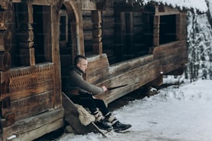 Strong viking warrior sharpening his sword while sitting near ancient wooden castle, scandinavian knight with weapon in viking costume, historical heritage concept