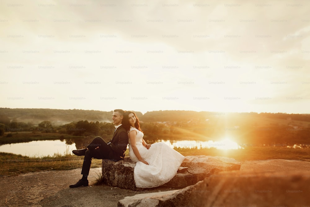 湖の風景の近くの夕日フィールドの石のベンチで休んでいる新婚夫婦のロマンチックなカップル、屋外で笑顔の花婿にもたれかかっている幸せな花嫁
