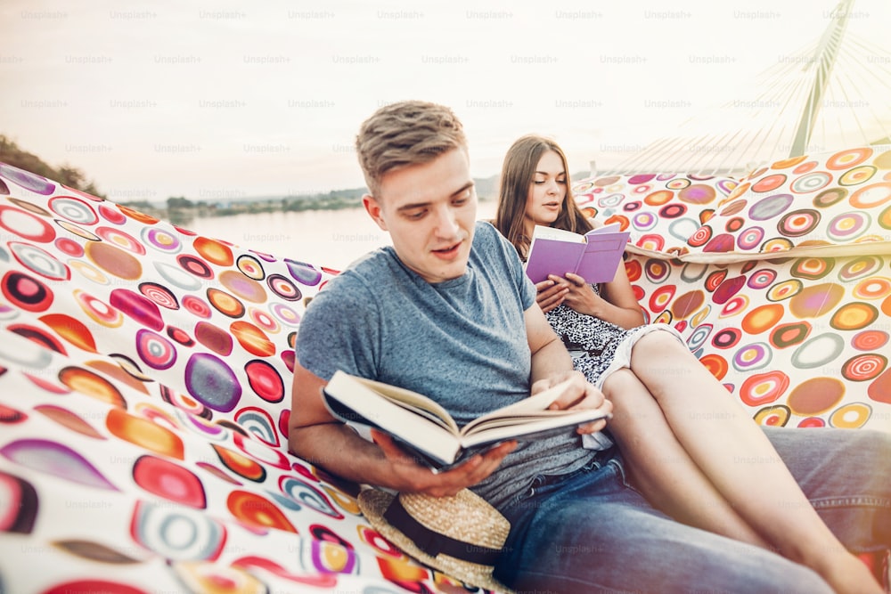 happy hipster couple reading books and relaxing in hammock in sunset light on the beach, summer vacation. stylish hipster family holding books and resting on holidays  in warm summer evening