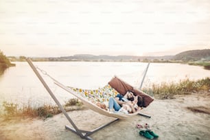 happy hipster couple with bulldog relaxing in hammock on the beach in sunset light, summer vacation. stylish family with dog cuddling and having fun, cute moments in summer evening