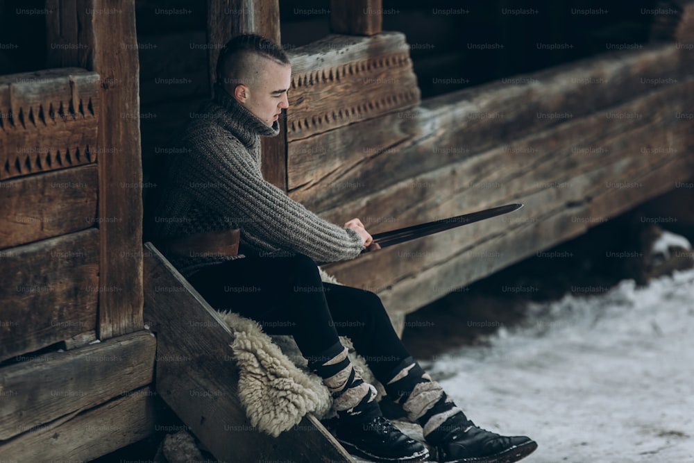 Starker Wikingerkrieger, der sein Schwert schärft, während er in der Nähe einer alten Holzburg sitzt, skandinavischer Ritter mit Waffe im Wikingerkostüm, historisches Kulturerbekonzept